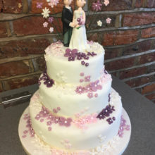 A white wedding cake with pink and purple flowers and icing cake toppers. The cake is on display in a shop in Wolverhampton.