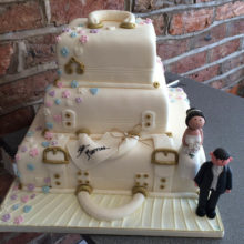 A three tiered wedding cake with each layer resembling a suitcase. The wedding cake includes a icing bride and groom.