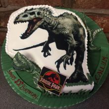 Personalised photo cake featuring a photo of a dinosaur. The cake is white and includes Jurassic Part icing stickers.