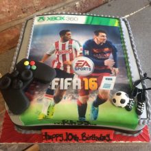 A personalised photo cake with a photo of a Fifa game on the front. The cake is square in shape and has decorative icing toppers.