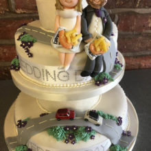 Three tiered wedding cake. The cake is white with two icing figures sitting on top. The cake has a icing road and cars.