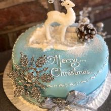 A bespoke cake, the cake is round and blue in colour. The cake says merry Christmas and has a icing reindeer on top.