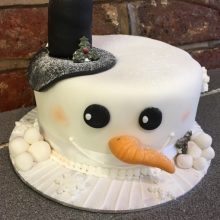 A bespoke cake crafted to look like a snowman. The snowman cake has a wonky nose with a top hat and black eyes.